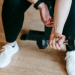 Closeup of lower half of woman wearing black workout leggings and white tennis shoes, who is seated and holding a dumbbell in one hand doing arm strength training exercises.