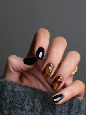 Photo courtesy of Canva Pro A close-up of the nail polish on a woman's left hand. Nails are active length with the thumb, pointer and pinky fingers painted in a glossy black polish while the middle and ring fingers are painted in a tortoiseshell finish of black and caramel brown.