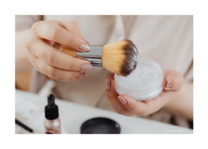 closeup of just a womans hands, with one hand holding a makeup brush and the other holding a container of sheer loose face powder.