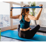 Midlife age woman wearing workout leggings and tank top sitting on exercise mat using a blue resistance band to do an upper back and shoulder strength training.