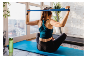 Woman wearing workout leggings and tank top, sitting on workout mat in living room, doing shoulder press using a resistance band.