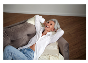 Smiling middle aged woman wearing jeans and a white button-up long sleeved top with the bottom buttons open showing a small part of her stomach, laying back with her arms behind her head on a living room couch.